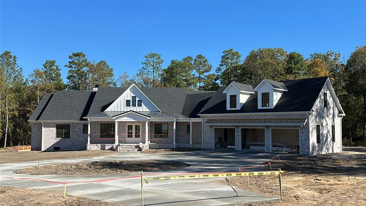 Large white custom residence under construction with brickwork, board-and-batten gables, dormers, porch columns, and three-car garage